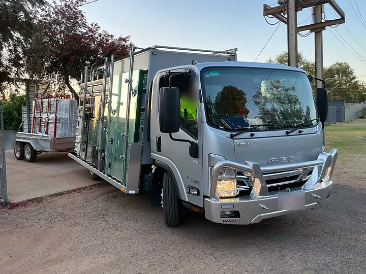 Silver Isuzu glass truck loaded with glass panels, with a trailer of glass behind it, at dusk
